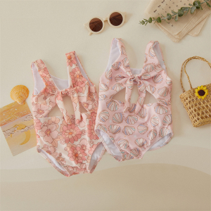 Two printed open-back one-piece baby swimsuits laid flat with sunglasses, a bag, and beach accessories