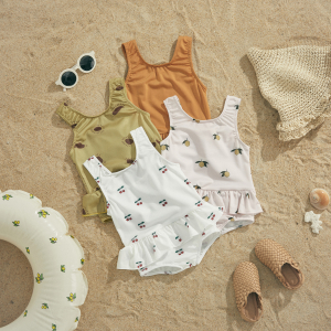 Four frilled one-piece baby swimsuits laid on sand with decorative beach accessories around them